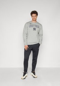 Grå hettegenser med "NEW YORK" trykket i marineblå, paret med svarte joggebukser. Skoene har en blanding av hvit og svart med oransje detaljer.