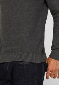 Maglione lavorato a maglia grigio scuro con orlo e polsini a coste, caratterizzato da un motivo testurizzato e un'etichetta laterale. Indossato sopra un paio di jeans in denim blu.