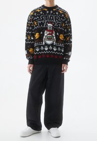 Schwarzer Strickpullover mit einem bunten "Star Wars"-Motiv, das Charaktere und Muster zeigt, kombiniert mit weiten schwarzen Hosen und weißen Sneakern.