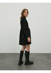 Zwarte jurk met lange mouwen, een getailleerde bodice en een uitlopende rok, gecombineerd met zwarte laarzen tot net boven de knie met een gestructureerde afwerking en dikke zolen.