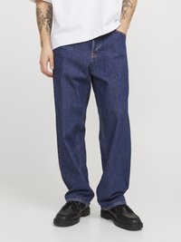 Jean en denim bleu foncé avec une coupe ample, dotée de coutures contrastées, de poches latérales et d'un ourlet retroussé. Porté avec des chaussures noires.