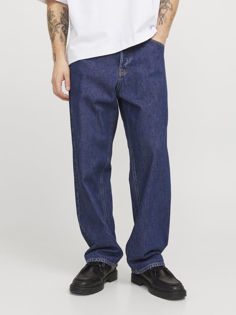 Jean en denim bleu foncé avec une coupe ample, dotée de coutures contrastées, de poches latérales et d'un ourlet retroussé. Porté avec des chaussures noires.