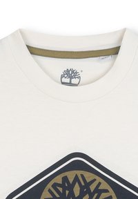Crèmekleurig katoenen t-shirt met een ronde halslijn en een gedrukt logo in donkerblauw en goud. Heeft een ribbelkraag en een gladde textuur.