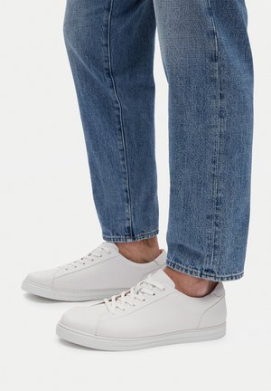 Person trägt blaue Jeans und weiße Low-Top-Schnürsenkel-Sneaker und steht auf einer weißen Oberfläche.