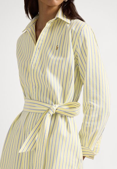 Kvinne iført en gul og lyseblå vertikalt stripet skjortekjole med belte knytt rundt livet og en liten brodert logo på brystet.