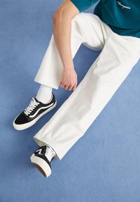 Zapatillas bajas en blanco y negro, con material de ante y lona, que presentan un diseño clásico de olas, combinadas con pantalones blancos sobre una superficie azul.