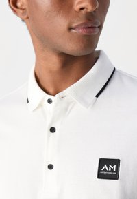 Polo bianco con colletto a costine, tre bottoni neri e un'etichetta quadrata nera con la scritta "AM" sul lato sinistro.
