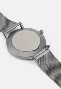 Grå rund klocka med mesharmband, med en slät metallbaksida och minimalistisk design. Minimal hårdvara med en ren finish.