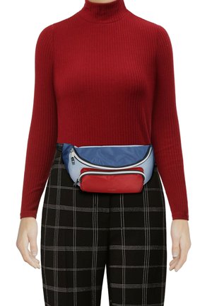 Maglione a collo alto rosso a coste, pantaloni a scacchi neri e una cintura a tracolla multicolore realizzata in materiali blu, rossi e bianchi con dettagli in cerniera.