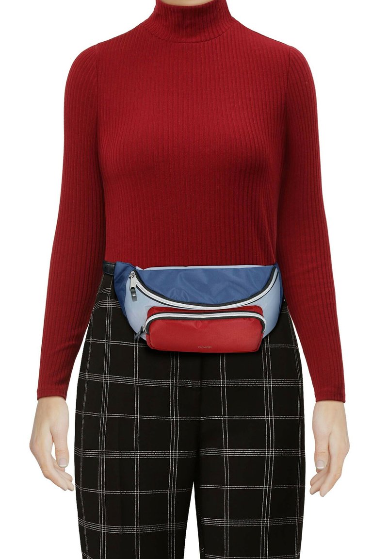 Maglione a collo alto rosso a coste, pantaloni a scacchi neri e una cintura a tracolla multicolore realizzata in materiali blu, rossi e bianchi con dettagli in cerniera.