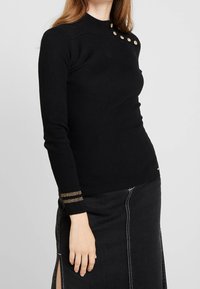 Pull noir côtelé à manches longues avec col montant, présentant des boutons à accent doré sur une épaule et un détail rayé doré sur les poignets.
