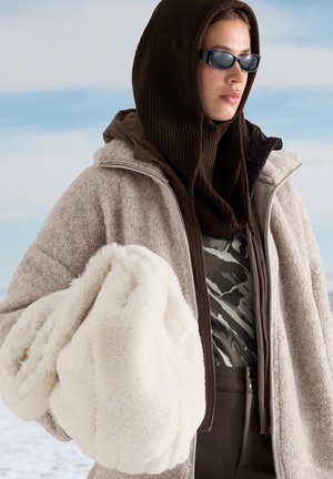 Hellbraune Fleecejacke mit Reißverschluss, darüber eine braun gemusterte Bluse. Weiße, flauschige Tasche, die unter einem Arm gehalten wird. Strukturierter brauner Schal.