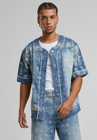 Denim shirt featuring a paisley pattern in various shades of blue, short sleeves, snap-button closure, and contrasting white stitching.