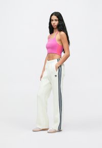 Mulher de pé com a mão no bolso, vestindo um top cropped rosa e calças de treino brancas da Adidas com riscas pretas, num fundo branco.