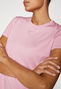 Světle růžové sportovní tričko s krátkým rukávem z hladké tkaniny, s jemným fialovým logem Nike u výstřihu a klasickým kulatým střihem.