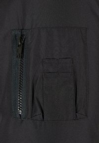 Detalle de una prenda de tela negra que presenta un bolsillo con cremallera con textura y un bolsillo de parche rectangular con costuras verticales.