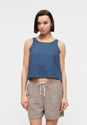 Žena s krátkymi hnedými vlasmi, oblečená v modrom tielku bez rukávov a béžových károvaných šortkách s šnúrkou, stojaca na jednoduchom šedom pozadí.