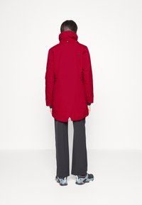 Chaqueta exterior roja con un collar alto, que presenta una abertura trasera y detalles del logo. Combinada con pantalones oscuros y zapatillas grisazuladas. Diseño minimalista.