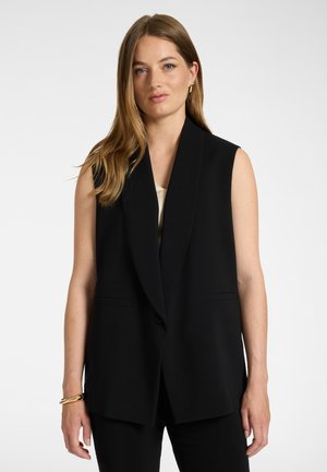 Donna con lunghi capelli castano chiaro che indossa un blazer nero senza maniche, orecchini a cerchio dorati e un braccialetto d'oro, in piedi davanti a uno sfondo semplice.