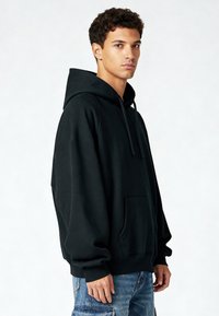 Zwarte hoodie met een trekkoord, grote voorkantzak en geribbelde manchetten. Draagt bij blauwe versleten jeans die een ontspannen pasvorm tonen.