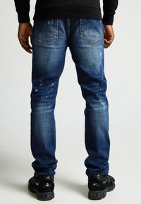 Jeans de mezclilla azul oscuro con ajuste slim, que presentan detalles desgastados y manchas de pintura ligera en la parte trasera. Bolsillos traseros lisos y costuras rematadas.