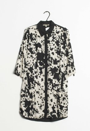 Chemise à motifs noir et blanc avec col, manches trois quarts, et poches avant. Tissu lisse avec une coupe ample et un ourlet arrondi.