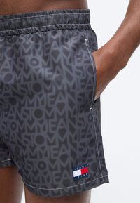 Άνδρας που φοράει σκούρα γκρι μαγιό με επαναλαμβανόμενο μοτίβο κειμένου "TOMMY JEANS" και πατζάκι με λογότυπο κοντά στο τελείωμα, με το χέρι στην τσέπη στο πλάι.