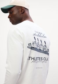 Dlouhý rukáv bílého trička s grafikou na zadní straně, na které jsou čtyři postavy v lodi a text v různých písmových stylech.