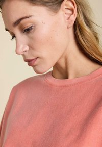 Sweat-shirt corail avec un col rond et une texture douce, présentant des variations subtiles de couleur et un minimum de détails, porté de manière décontractée.