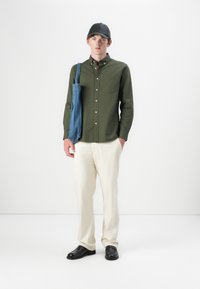 Camicia verde a bottoni, pantaloni crema, scarpe nere e una borsa in denim blu. La camicia presenta una tasca sul petto e un colletto.