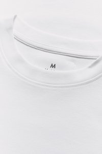 Vit bomullst-shirt med rund halsringning och synliga sömmar. Etiketten visar storlek medium. Slät textur, utan mönster eller grafik.