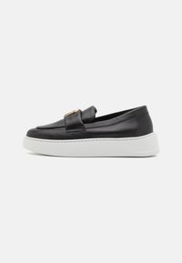 Zapato negro de piel sin cordones con una textura suave, que presenta un acento dorado en la correa y una suela de plataforma blanca. Vista lateral mostrada.
