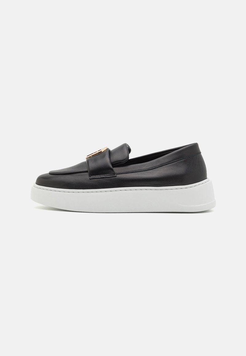 Zapato negro de piel sin cordones con una textura suave, que presenta un acento dorado en la correa y una suela de plataforma blanca. Vista lateral mostrada.