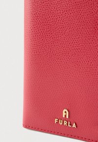 Κόκκινο πορτοφόλι από υφασμάτινο δέρμα με χρυσό λογότυπο. Διαθέτει ορατή ραφή και κομψό σχέδιο. Συμπαγές και ορθογώνιο σχήμα.