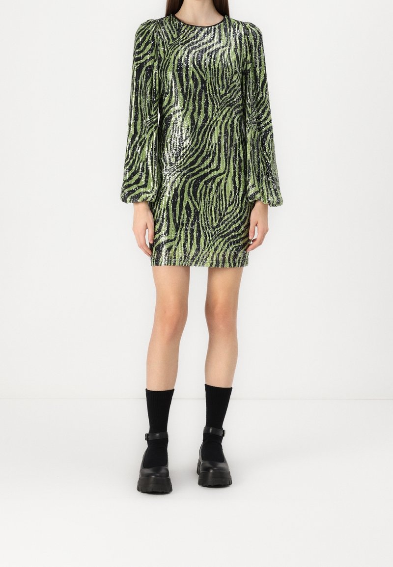 Femme portant une mini-robe à paillettes imprimé zèbre vert et noir, avec des manches longues bouffantes, des chaussettes noires et des chaussures noires épaisses à bride.
