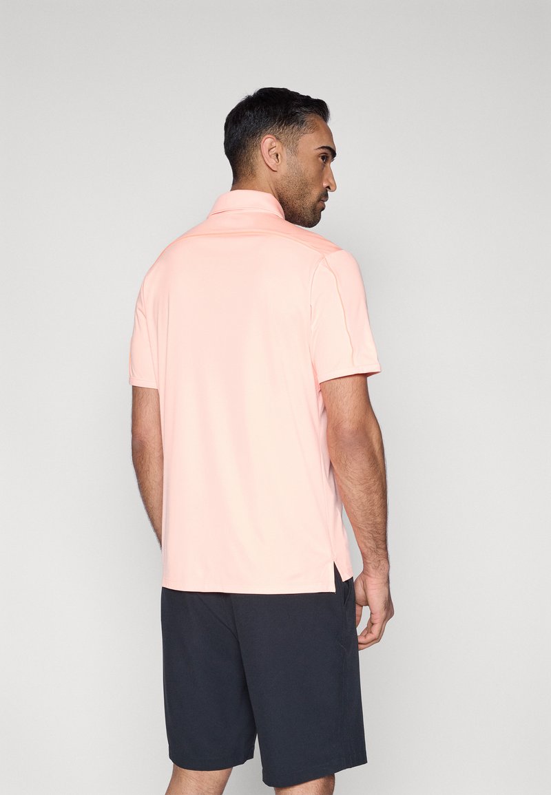Polo pour homme à manches courtes de couleur rose clair, fabriqué en tissu lisse avec un col, associé à un short foncé présentant des fentes latérales.