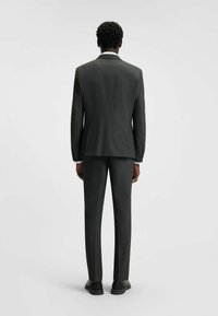 Dunkelgrauer Anzug mit subtiler Textur, maßgeschneidertem Design, einem Schlitz im Rücken und slim-fit Hosen. Schwarze Schuhe ergänzen das Outfit.