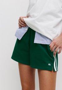 Grüne Baumwollshorts mit weißen Seitenstreifen und einem kleinen Logo am Saum. Kombiniert mit einem weiß-blauen, gestreiften Hemd mit Knöpfen.