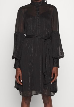 Femme portant une robe noire transparente à manches longues avec boutons, col montant, ceinture à la taille et discrètes rayures verticales scintillantes.