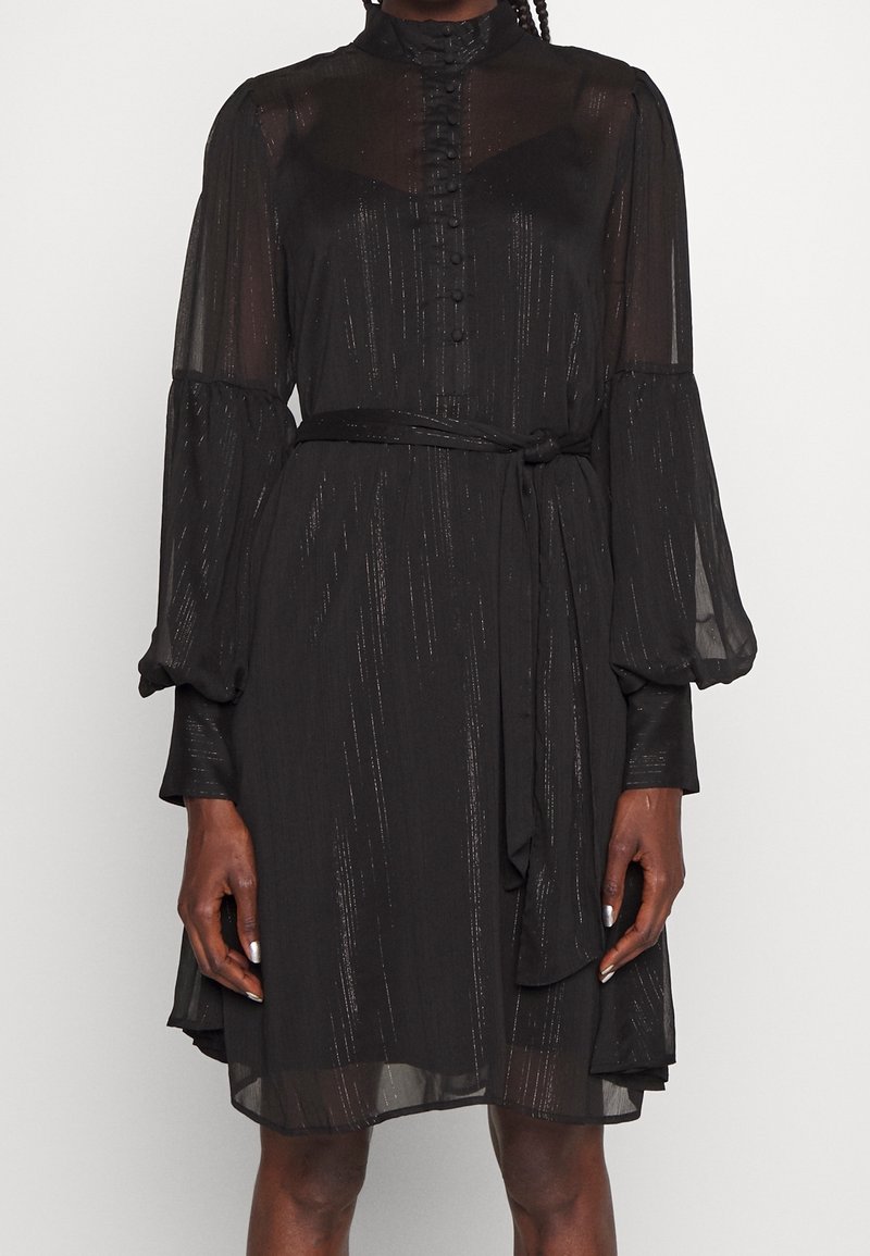 Femme portant une robe noire transparente à manches longues avec boutons, col montant, ceinture à la taille et discrètes rayures verticales scintillantes.