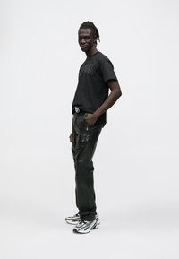 Muž s vrkočmi stojí bokom, má na sebe čierne tričko s potlačou, čierne cargo nohavice a bielo-čierne tenisky na jednofarebnom pozadí.