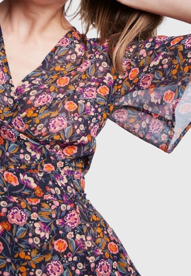 Blusa floreale con sfondo scuro, caratterizzata da fiori vivaci in tonalità di viola, arancione e rosa. Maniche trasparenti e fluenti con design avvolgente.