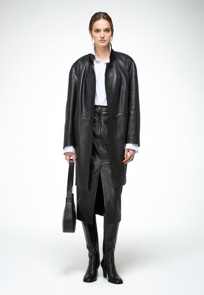 Frau trägt einen schwarzen Lederjacke, passenden Rock mit vorderem Schlitz, schwarze Stiefel mit Absatz, weißes Hemd und trägt eine schwarze Handtasche.