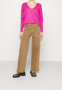 Suéter de punto rosa brillante con escote en V, combinado con pantalones beige de talle alto y pierna ancha y zapatos de tacón bronce. Diseño sencillo.