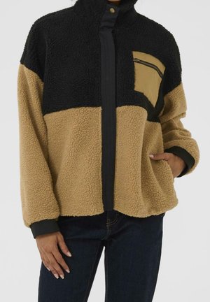 Veste polaire bicolore beige et noire avec fermeture éclair et poche zippée en biais, portée avec un jean foncé, mains dans les poches, sur fond neutre.