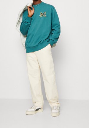 Mężczyzna w turkusowej bluzie z żółtym logo "M", kremowych spodniach, białych trampkach, trzyma lekka kurtkę zarzuconą na ramię na białym tle.