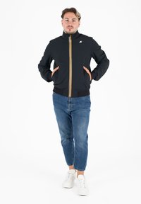 Zwarte bomberjack met gele en oranje rits, geribde manchetten en zoom, gedragen met blauwe jeans en witte sneakers, handen in de zakken.