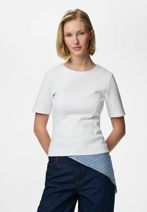 Femme aux cheveux blonds courts portant un haut blanc à manches courtes superposé à une chemise rayée bleue et un jean bleu foncé, sur un fond uni.
