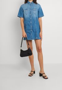 Vestido camisa de mezclilla con mangas cortas, cierre de botones, dos bolsillos en el pecho y un lavado en azul claro. Accesorios incluyen un bolso negro y tacones con tiras.