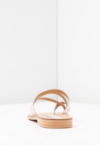 Enkel tan flad sandal med to tynde læderstropper, set fra hælen på en hvid overflade og hvid baggrund.